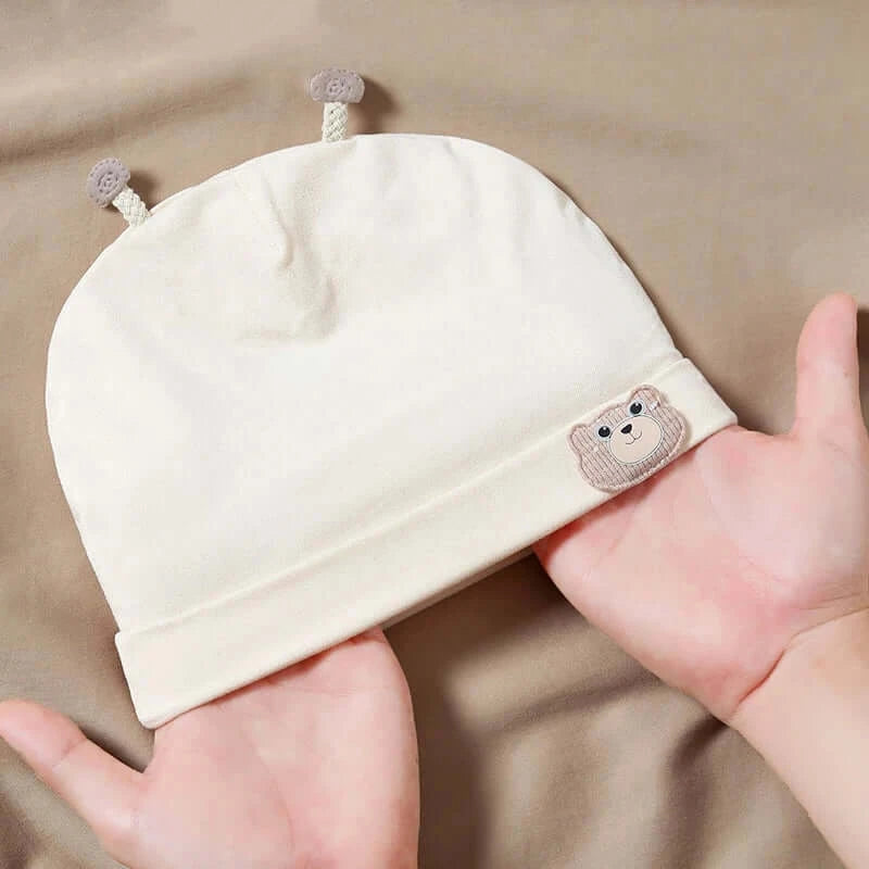 Cream-colored baby beanie with bear patch, soft cotton fabric, designed for newborns, held in hands for size comparison