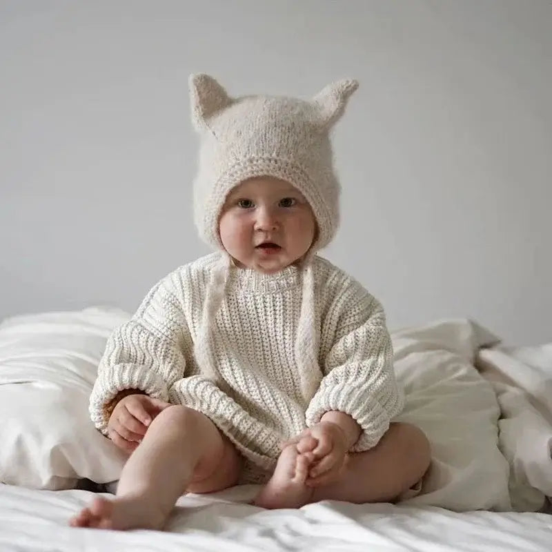 Baby Beanie mit Hasenohren auf einem süßen Baby in einem gestrickten Outfit, das auf einem Bett sitzt.